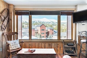 A room with a large window, a wooden chair with a cushion, a table with books, and a television mounted on the wall. - Peak 9 Inn 4512: Stellar Views - Downtown & Ski In Ski Out @ Base of Peak 9 (Breckenridge)