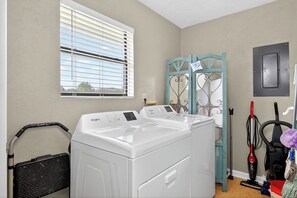 A laundry room with two front-loading washing machines, a window with blinds, a blue metal gate, and a vacuum cleaner. - Key Lime Cottage - 250 Steps to Beach, Beach Gear Credit Included, Recent Renovations (St. George Island)