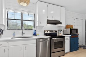A modern kitchen with white cabinetry, stainless steel appliances, and a window with blinds. - Key Lime Cottage - 250 Steps to Beach, Beach Gear Credit Included, Recent Renovations (St. George Island)