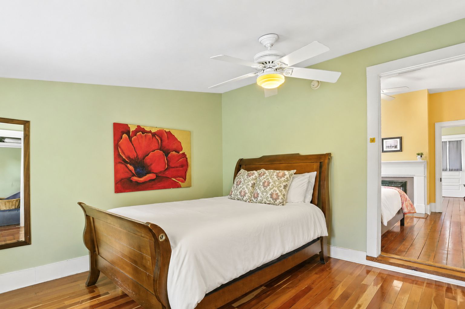 Quarto com cama de madeira, ventilador de teto, pintura de uma flor vermelha e vista para um corredor.
