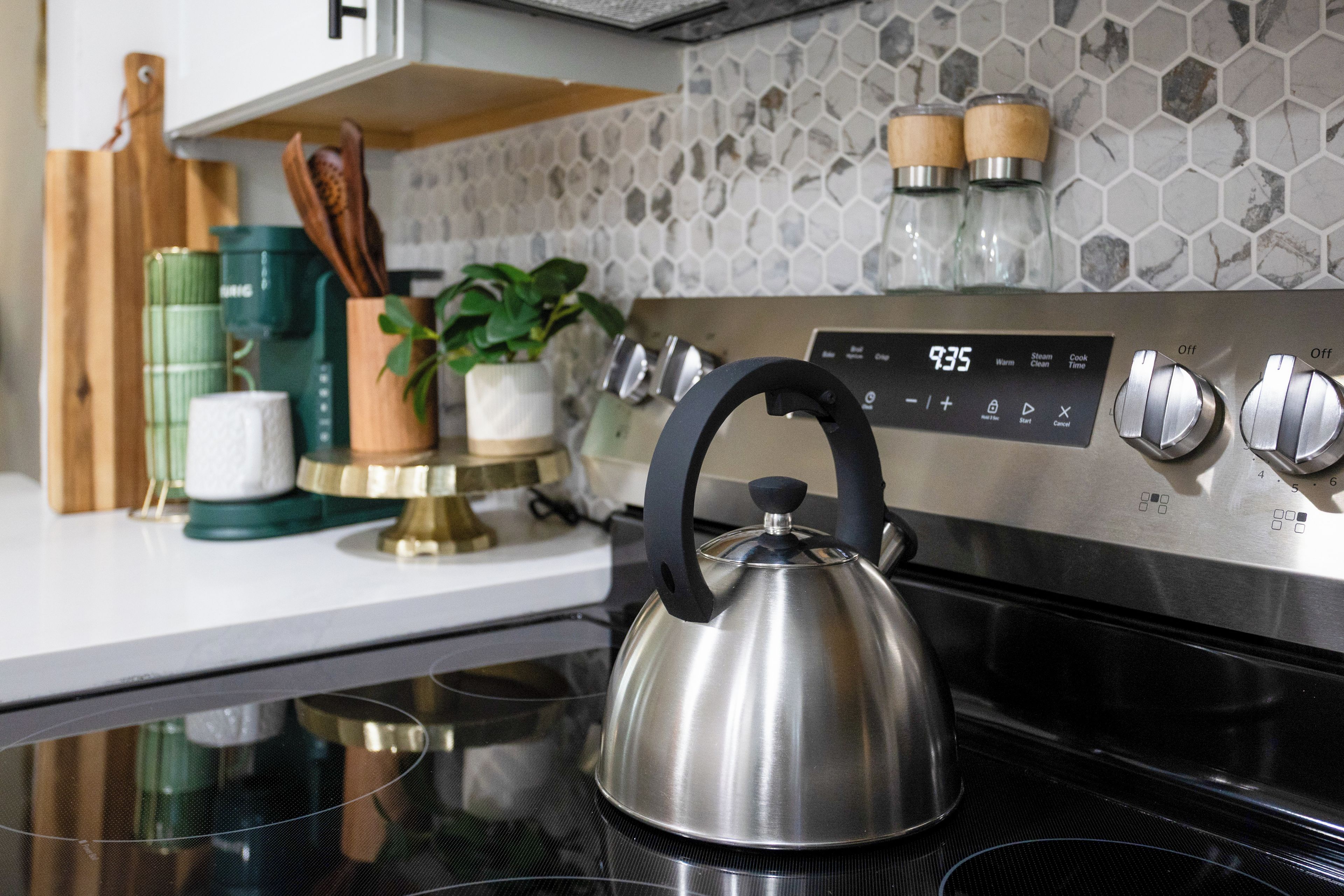 A stainless steel kettle on a black stovetop, a digital clock displaying 7:35, and a kitchen counter with various items including a green container, a white cup, and wooden utensils.