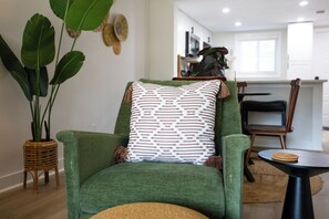 A green armchair with a patterned pillow, a woven side table, and a large potted plant.