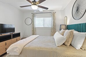 A bedroom with a bed, a ceiling fan, a TV, a window with curtains, and a wall art piece.