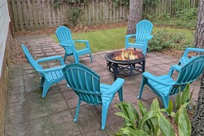 Eine Terrasse mit vier blauen Stühlen, die um einen runden Tisch mit einem Feuerkorb in der Mitte angeordnet sind.