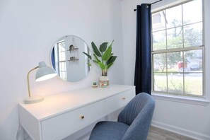 Een kamer met een witte ladekast, een ronde spiegel, een bureaulamp, een plant in pot en een raam met gordijnen.