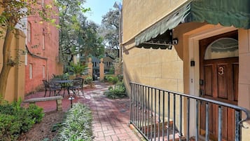 A courtyard with a brick pathway, outdoor seating, and a building entrance with a door and window.