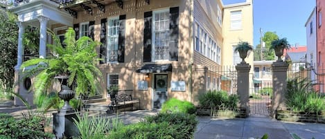 A two-story building with a balcony, a covered entrance, and a garden with a fountain.