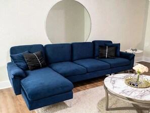 Ein blaues Ecksofa mit Chaiselongue, ein runder Marmortisch und ein Beistelltisch mit Gläsern und einer Blumenvase.