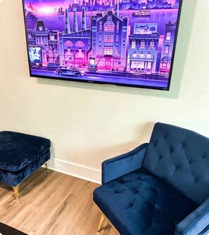Ein Zimmer mit einem großen Flachbildfernseher an der Wand, der eine Stadtansicht zeigt. Zwei dunkelblaue, gepolsterte Sessel mit Wirbelrückseiten.