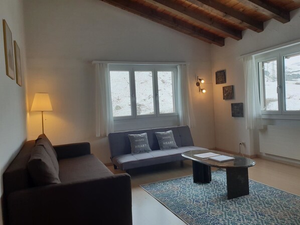 A living room with a sofa, a coffee table, and a lamp. There are windows with curtains, a wall with framed pictures, and a ceiling with exposed beams.