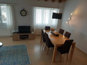 A modern living room with a wooden dining table, black chairs, a flat-screen TV, a clock on the wall, and a large window with sheer curtains. - Curtinfess Franklin Appartement/Fewo, Bad, WC, 2 Schlafräume by Interhome (Bivio)