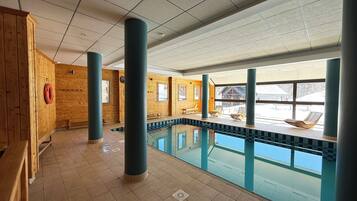 An indoor swimming pool with wooden walls, large pillars, and a view of a snowy landscape.