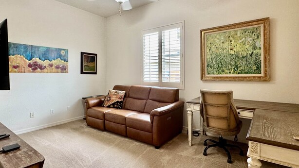 A room with a brown leather sofa, a wooden desk, a chair, a ceiling fan, and two paintings on the walls. - Oro Valley Home For Rent w/ Spa & Views (Oro Valley)