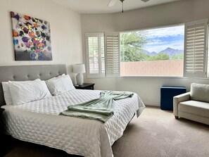 Ein Schlafzimmer mit Bett, Nachttisch, Lampe, einem Fenster mit Jalousien, Blick auf Grünflächen und Berge sowie einem Bild an der Wand.