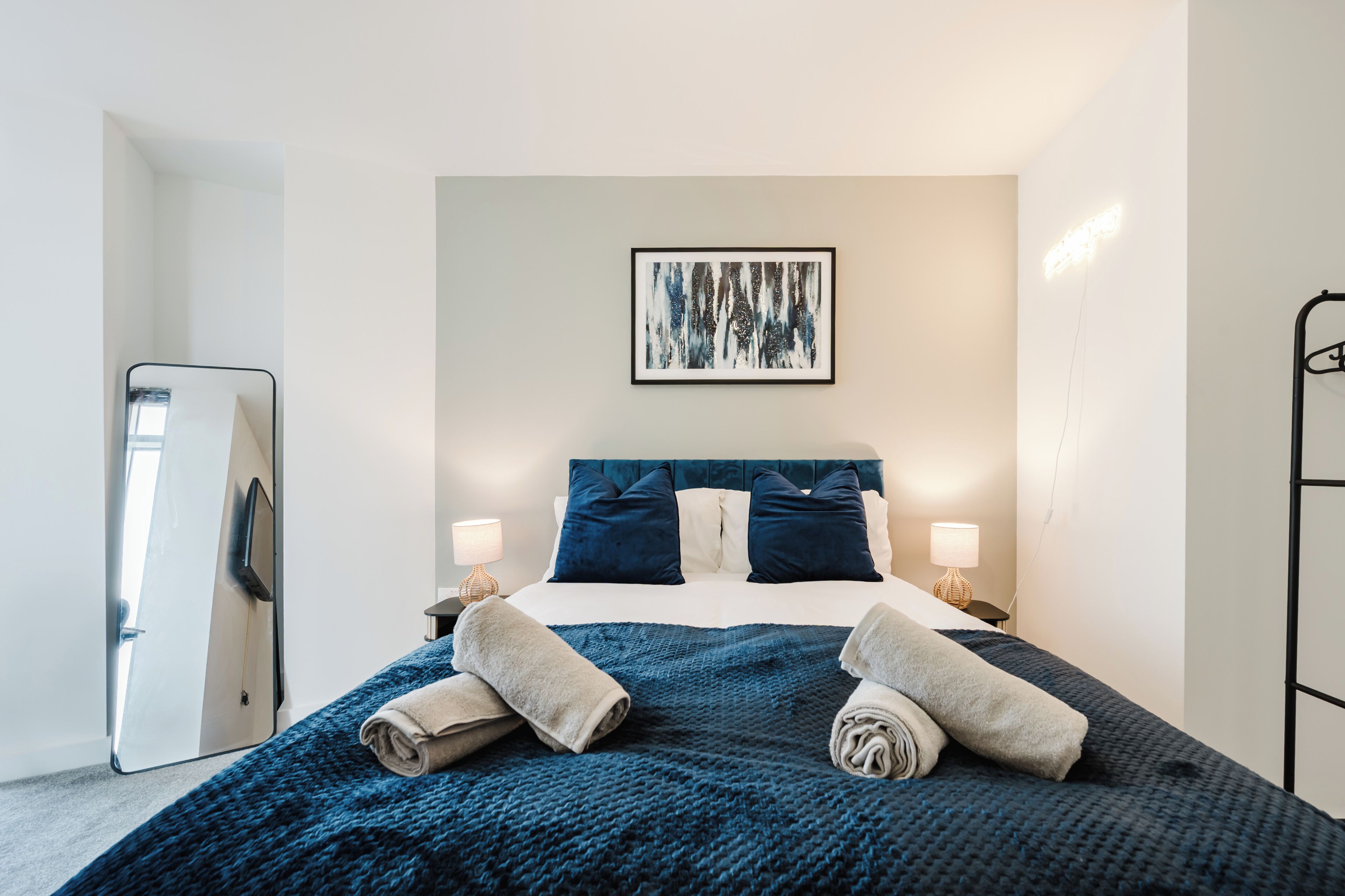A modern hotel room with a bed, two bedside lamps, a large mirror, and a framed abstract painting on the wall.