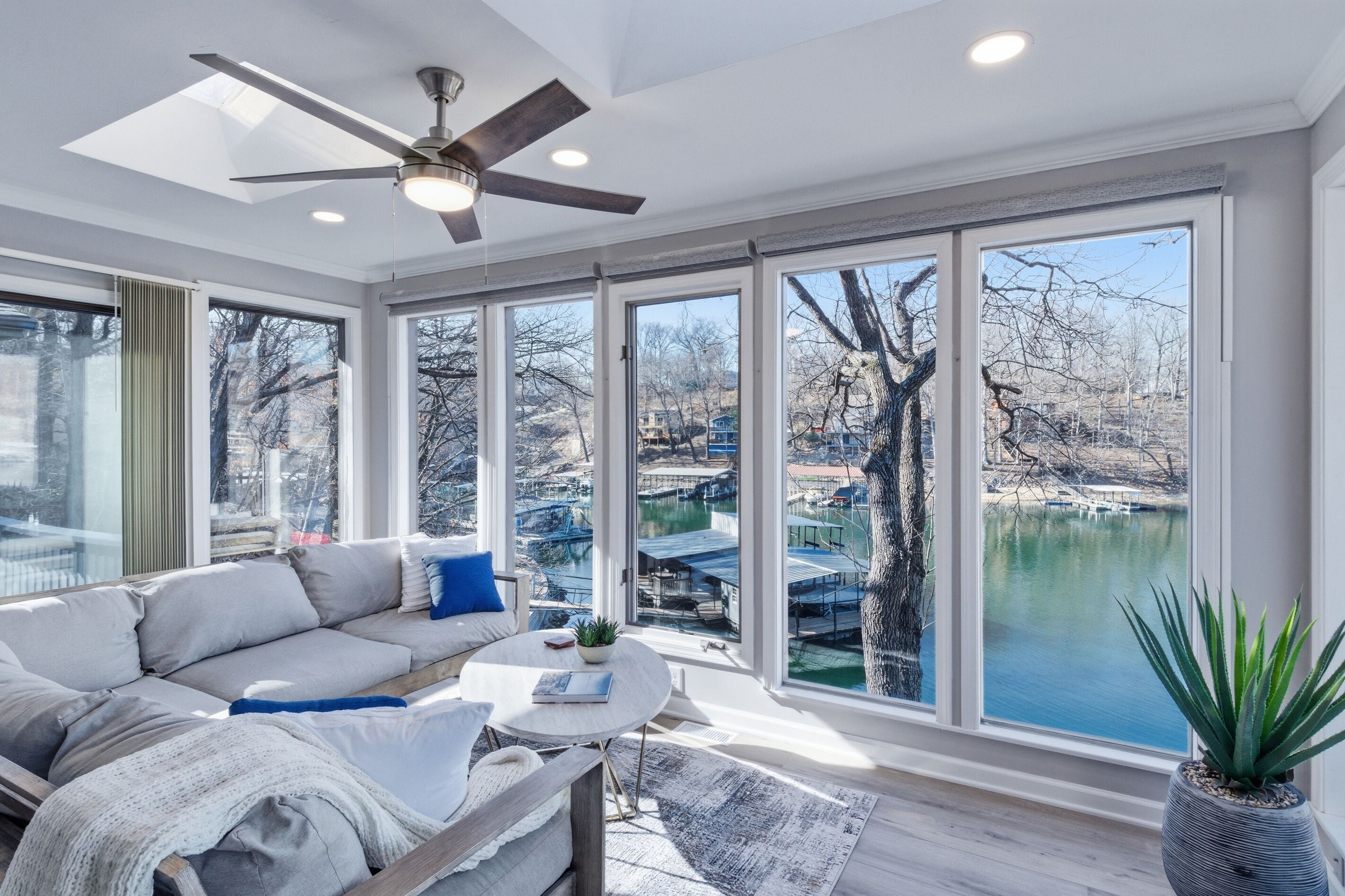 A modern living room with a sofa, coffee table, and a ceiling fan. Large windows offer a view of a body of water and buildings.