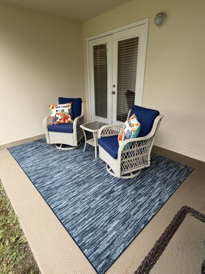 Eine Terrasse mit zwei Korbstühlen, einem kleinen Tisch und einem blauen Teppich.