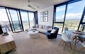 A spacious living room with a large window, a sofa, a coffee table, and a dining area with chairs. - Luxury Penthouse | Sandbar Burleigh Heads (Gold Coast)