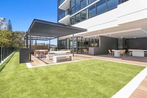 Un patio extérieur moderne doté d’un espace salon couvert, d’une pelouse et d’un bâtiment avec de grandes fenêtres et un toit plat.