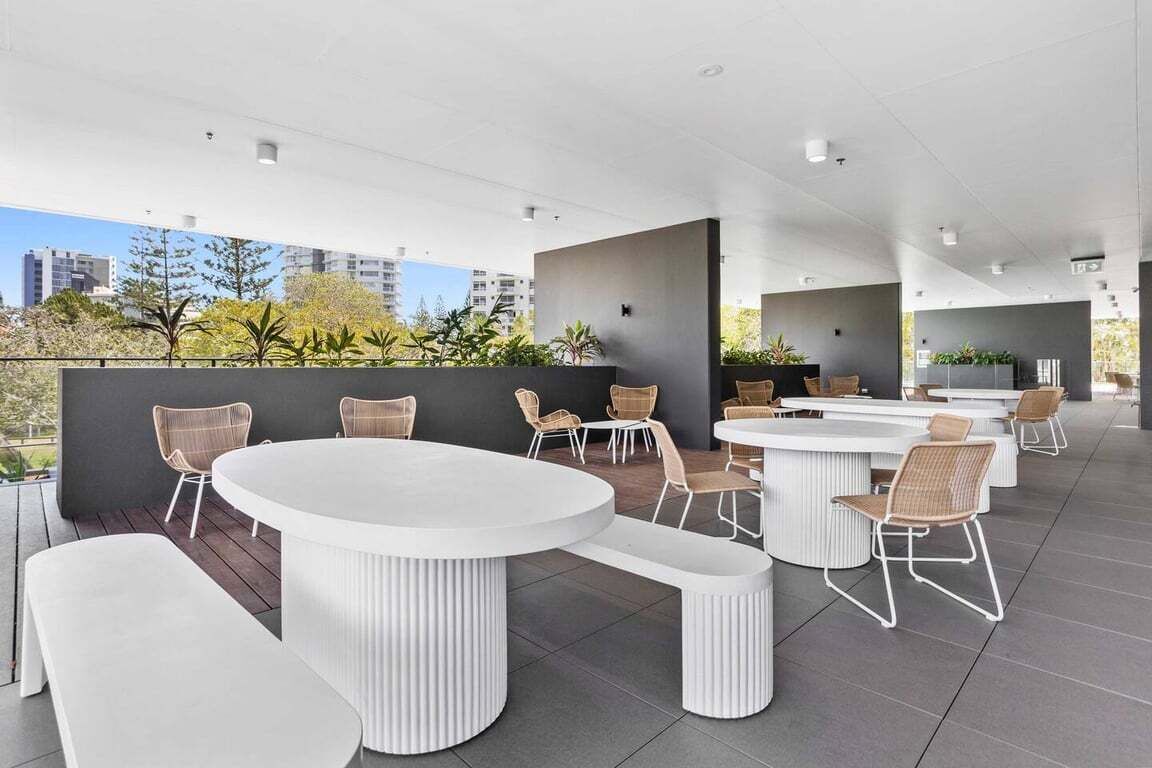 An outdoor seating area with round tables and wicker chairs, surrounded by a low wall and plants.