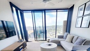 A modern living room with a large window, a ceiling fan, a sofa, a coffee table, and a TV. - Luxury Penthouse | Sandbar Burleigh Heads (Gold Coast)