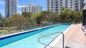 A swimming pool with a clear blue water, surrounded by a wooden deck and glass railing. - Luxury Penthouse | Sandbar Burleigh Heads (Gold Coast)