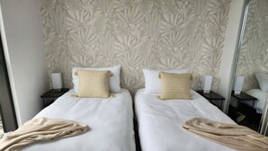 A hotel room with two beds, each with a white bedspread and a beige throw blanket. There are two bedside tables with lamps, and the walls have a leaf-patterned wallpaper. - Luxury Penthouse | Sandbar Burleigh Heads (Gold Coast)
