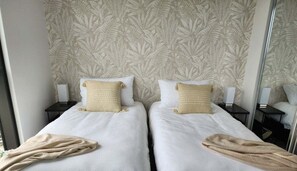 Ein Hotelzimmer mit zwei Betten, jeweils mit weißer Bettdecke und beige-farbener Tagesdecke. Zwei Nachttische mit Lampen stehen bereit, und die Wände sind mit einem Blattmuster tapeziert.