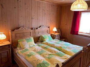 A wooden room with a double bed, two bedside tables, a hanging light fixture, and a window with red curtains. - Tga Crap Appartement/Fewo, Dusche, WC, 1 Schlafraum 2.5 Zimmerwohnung - 4 Betten by Interhome (Savognin)