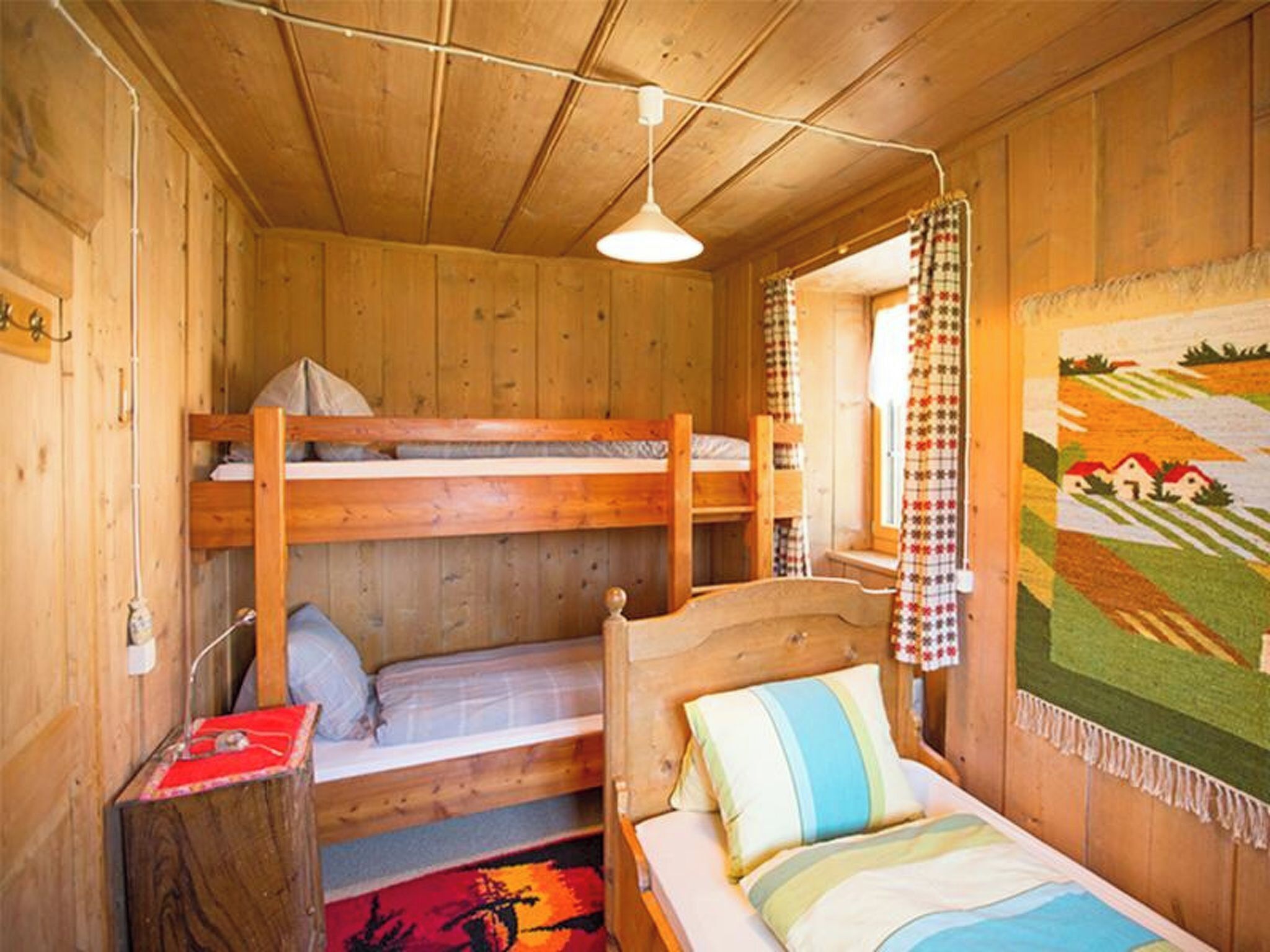 A wooden cabin room with bunk beds, a single bed, a hanging light fixture, and a tapestry on the wall.
