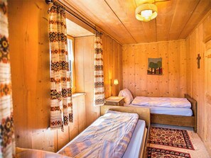 A wooden cabin room with two beds, a patterned curtain, a small table with a lamp, and a wall painting.
