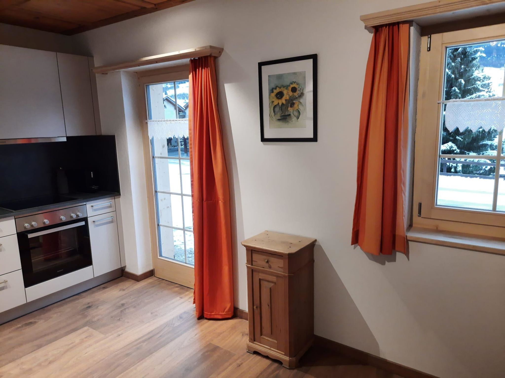 Une cuisine équipée d’un four encastré, d’un petit placard et d’une fenêtre ornée de rideaux orange.