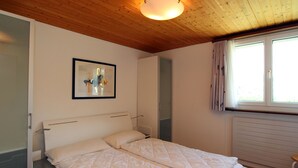 Un dormitorio con cama doble, mesita de noche, cuadro abstracto enmarco, ventana con cortinas y techo de madera.