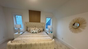 Ein Schlafzimmer mit einer gebügelten Kopfstütze, einem großen Bett, zwei Nachttischen mit Lampen, einem Fenster und einem an der Wand befestigten Spiegel.