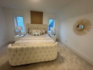 Een slaapkamer met een geborderde hoofdbord, een groot bed, twee nachtkastjes met lampen, een raam en een spiegel aan de muur.