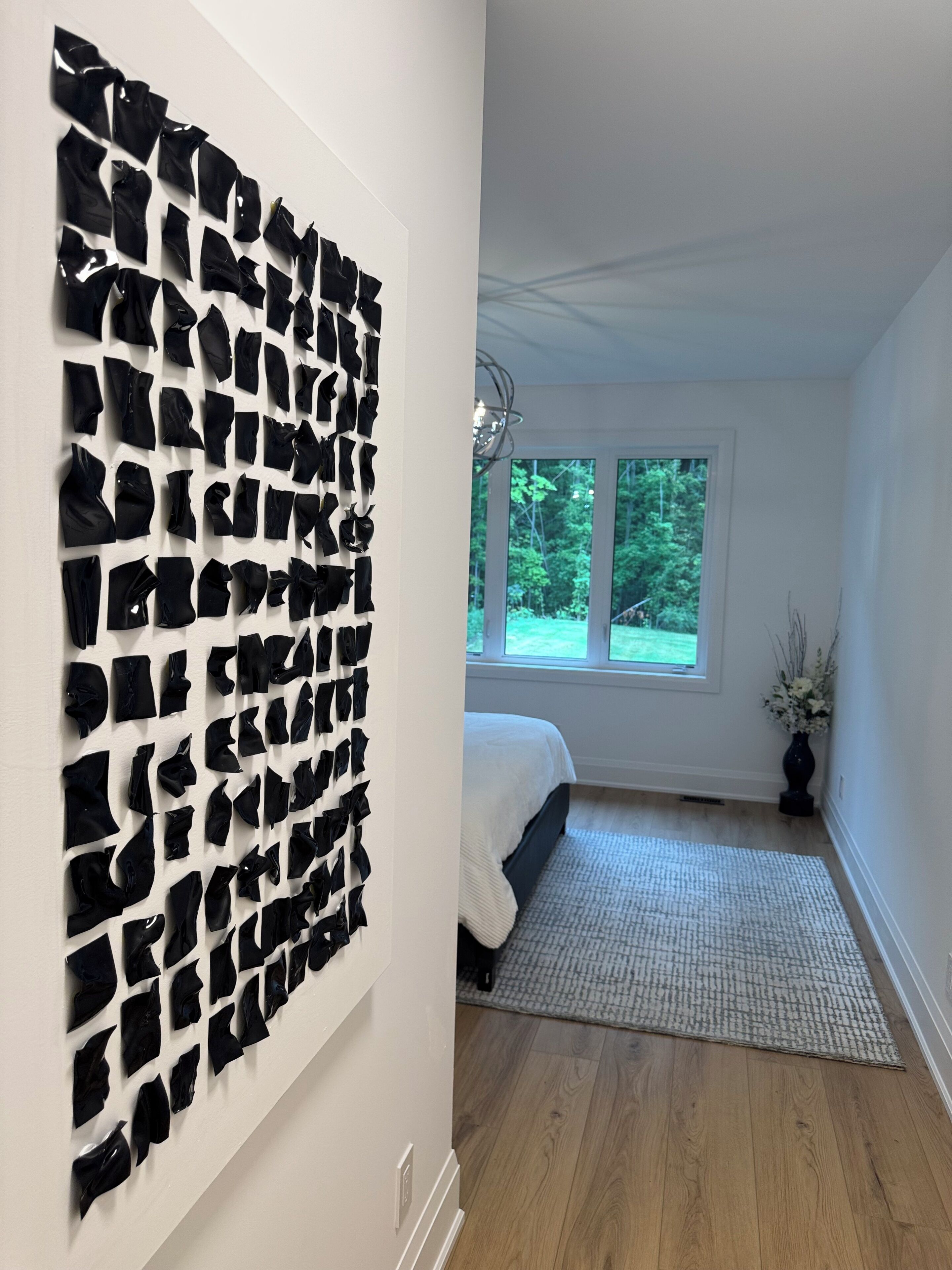 Une pièce dans laquelle on trouve une grande installation artistique abstraite au mur, un lit recouvert d’une couverture blanche et une fenêtre donnant sur des arbres.
