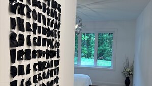 A room with a large abstract art installation on the wall, a bed with a white cover, and a window with a view of trees.