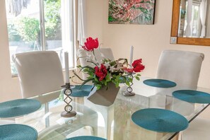 A dining table with a glass top, white chairs, and teal placemats. A vase with red and white flowers is on the table.