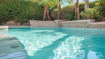Una piscina cristalina con una pared de piedra y escalones que bajan hacia ella, rodeada de exuberante vegetación y palmeras.