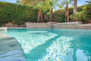 A clear pool with a stone wall and steps leading into it, surrounded by lush greenery and palm trees.
