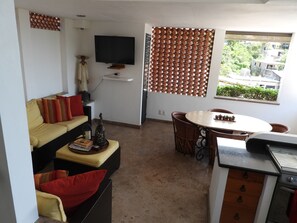Ein kompakter Wohnraum mit einer Couch, einem Esstisch mit Stühlen, einem an der Wand befestigten Fernseher und einem Fenster mit Blick auf Grünflächen.