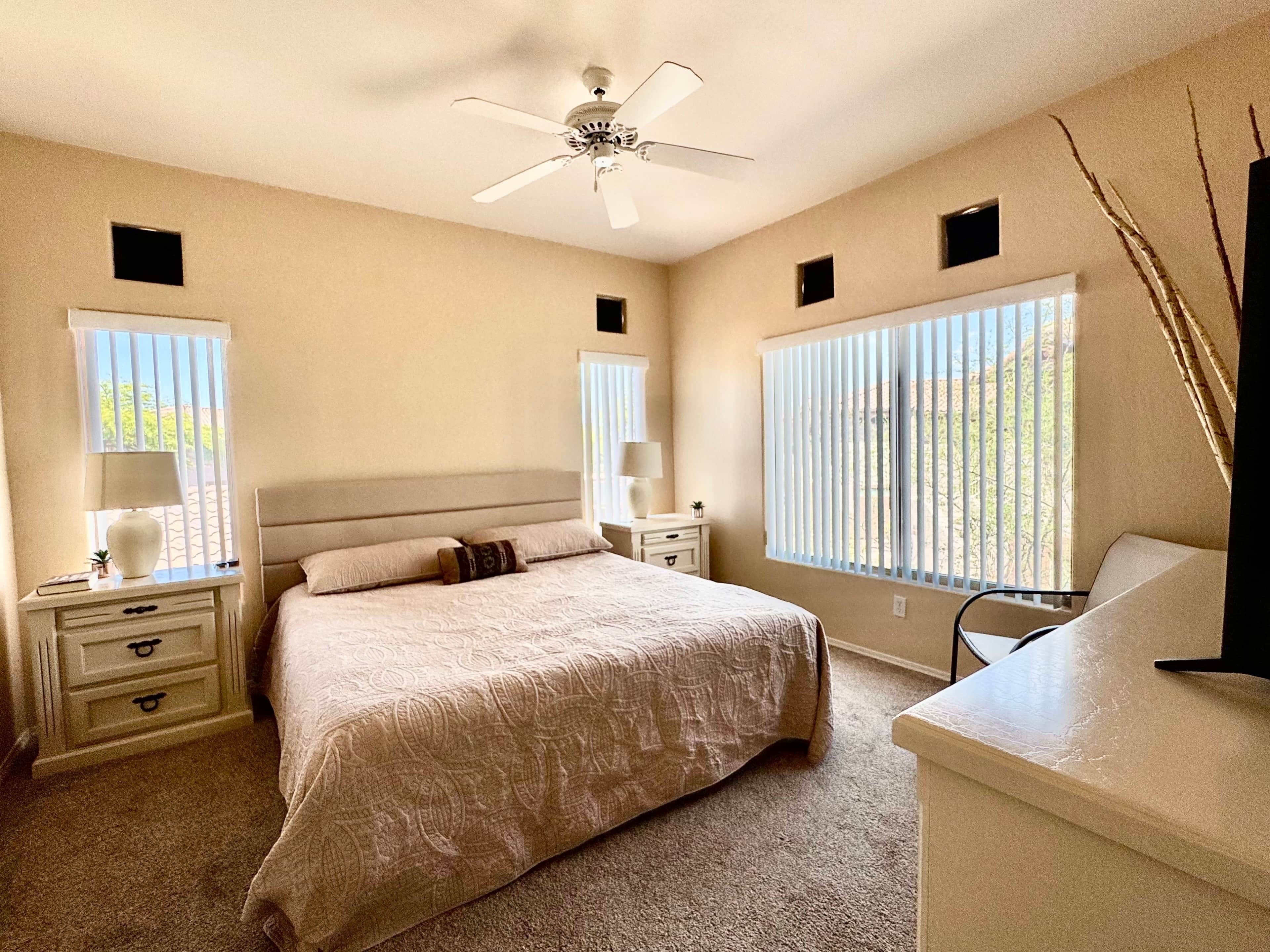 A bedroom with a bed, bedside tables, a ceiling fan, and windows with blinds.