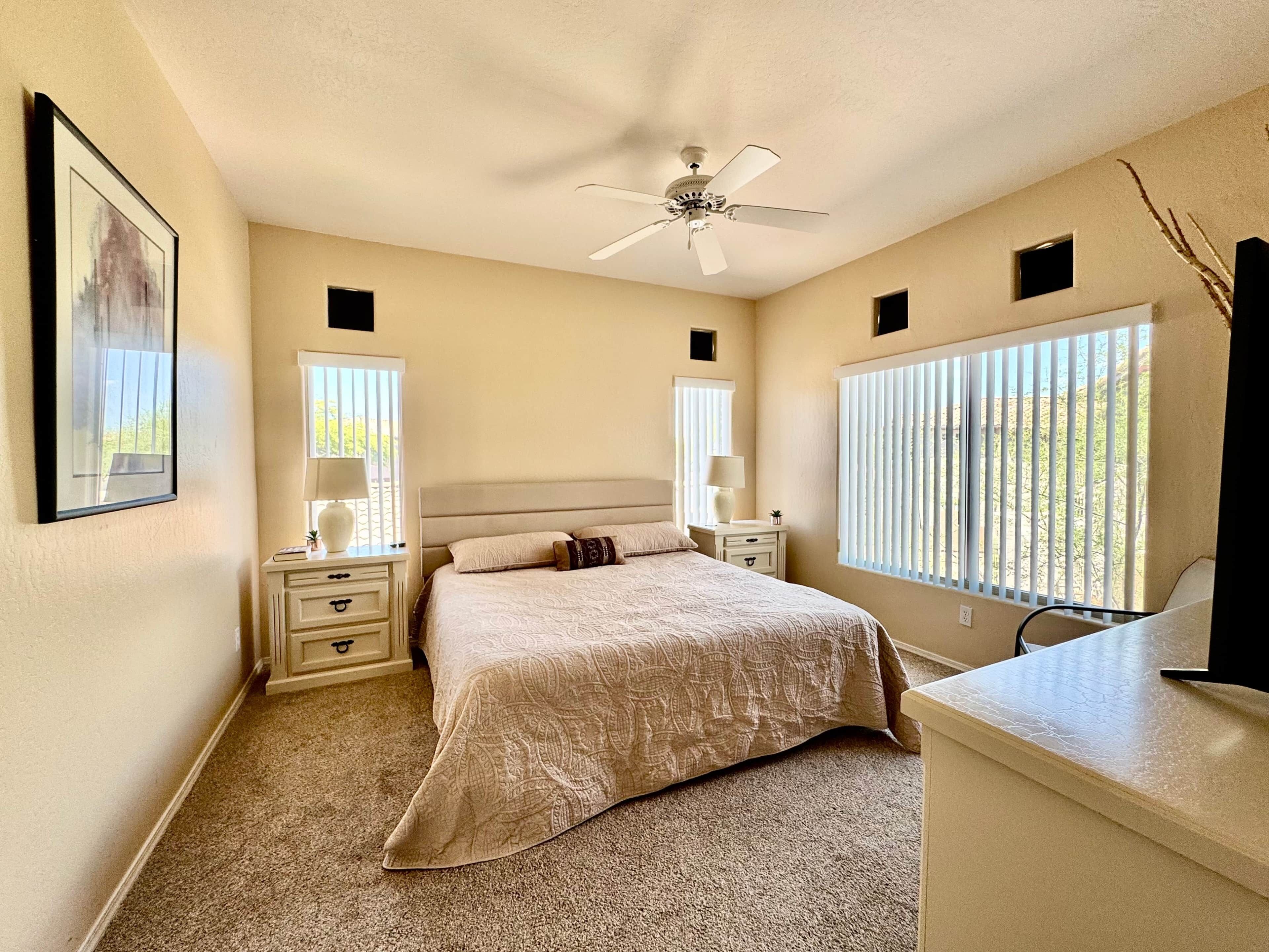 A bedroom with a bed, bedside tables, a ceiling fan, and windows with blinds.