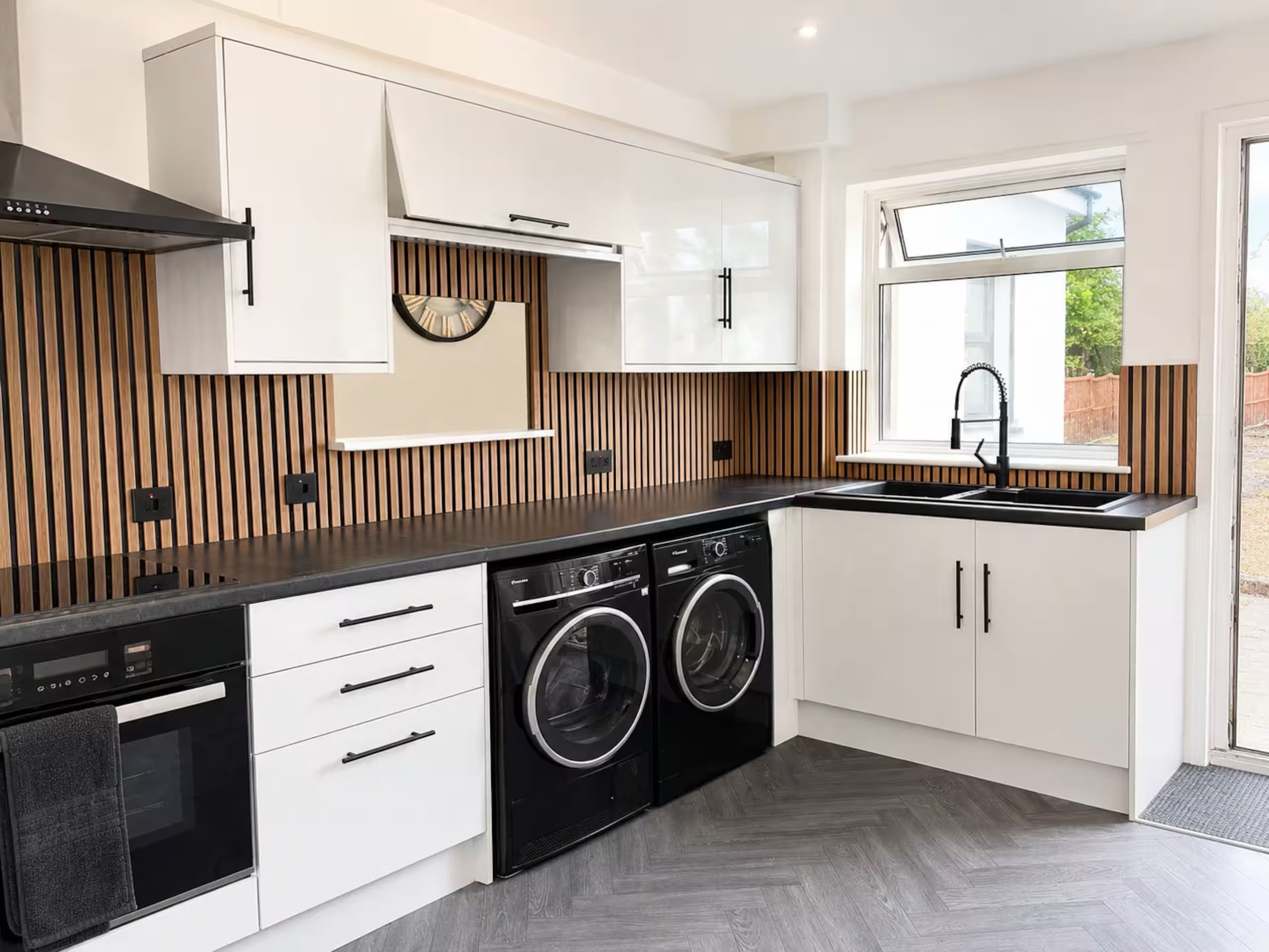 A modern kitchen with built-in appliances, a window, and a herringbone wood panel backsplash.