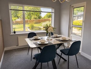 A dining area with a table set for four, two chairs on each side, a vase with flowers, and a view of a garden through the window. - Vivian - Luxury 2 bed House (Watford)