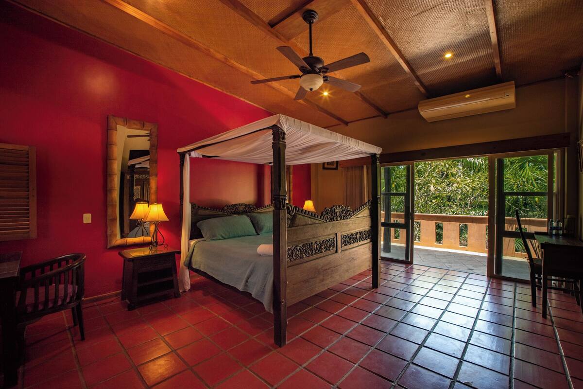 A four-poster bed with a canopy, a ceiling fan, and an air conditioning unit in a room with red walls and tiled flooring.