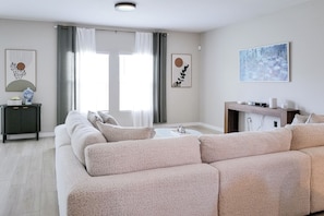 A modern living room with a beige sofa, a coffee table, and a fireplace. There are two windows with curtains, a painting on the wall, and a small side table with a vase. - Enjoy this Home in Wesley Chapel.  Designed for Relaxation/Comfort/Remote Work (Wesley Chapel)