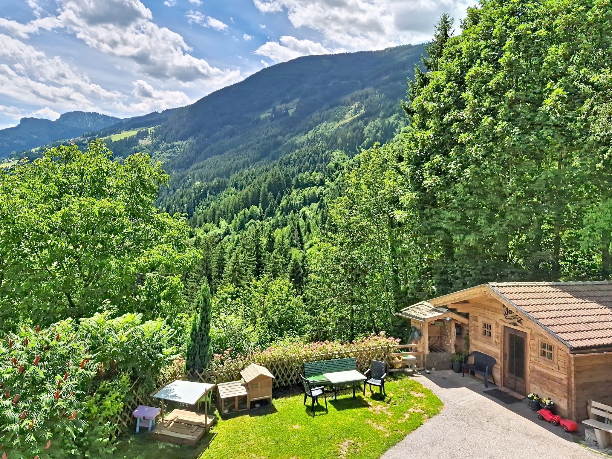 A wooden cabin with a patio area, surrounded by lush greenery and mountains in the background.