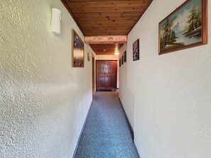 Ein Korridor mit Holzboden, weißen Wänden und Teppichboden. An den Wänden befinden sich Bilder in Rahmen, und am Ende des Ganges eine Tür.