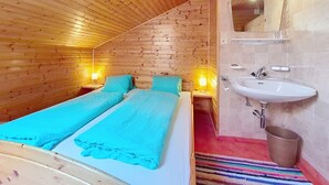 Um quarto com teto de madeira inclinado, cama com roupa de cama azul, pia branca e um espelho.